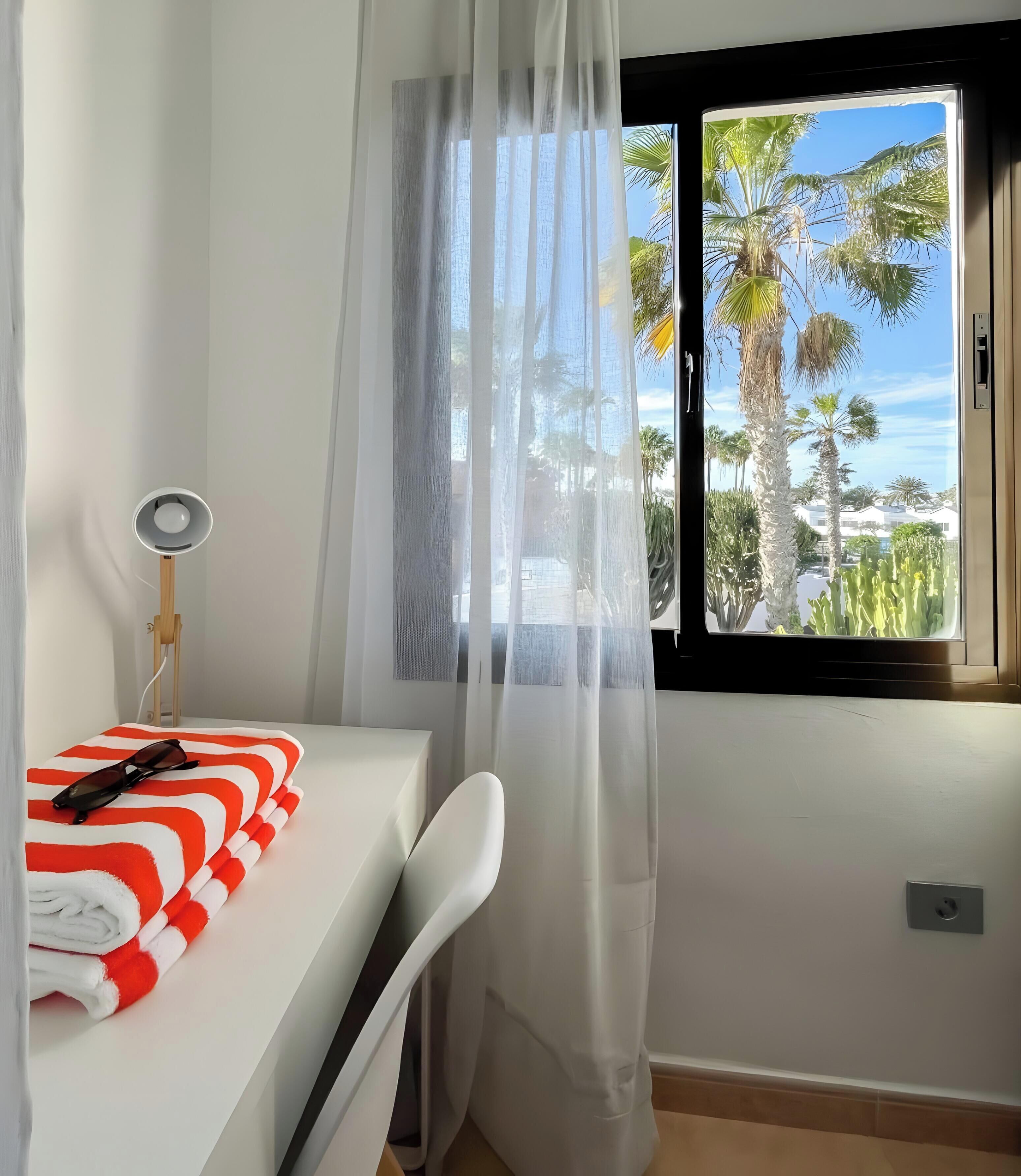 Un dormitorio con una cama, una mesita de noche con una lámpara, un ventanal con vista a palmeras y una cortina.