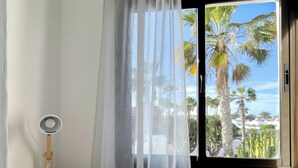 Une chambre à coucher avec un lit, une table de chevet avec une lampe, une fenêtre donnant sur des palmiers et un rideau.