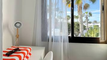 Un dormitorio con una cama, una mesita de noche con una lámpara, un ventanal con vista a palmeras y una cortina.