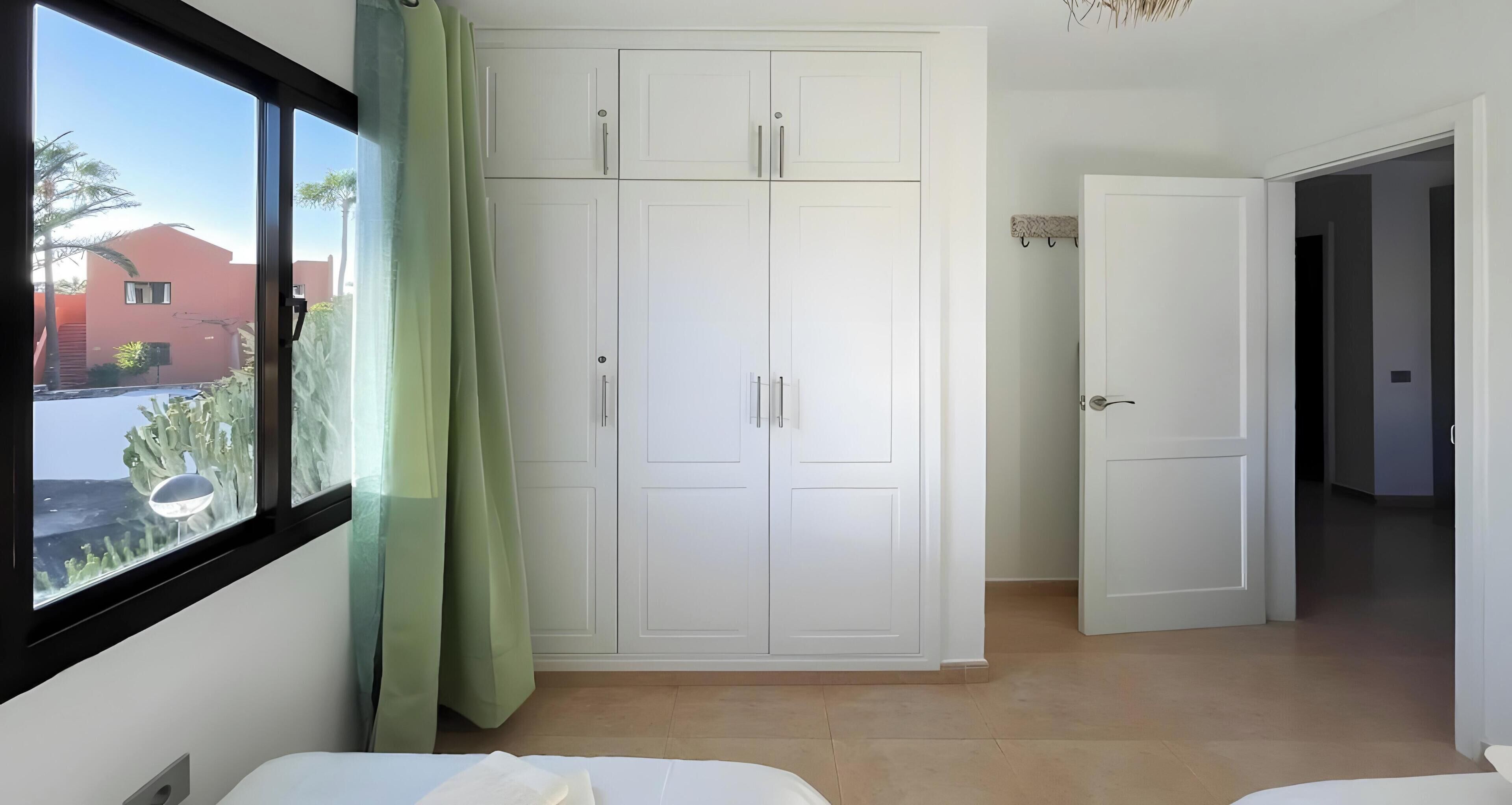 Une pièce avec une grande armoire blanche, une fenêtre donnant sur des palmiers et une porte menant à une autre pièce.