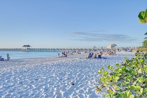 Ein Strand mit einem Steg, der sich ins Wasser erstreckt, Menschen entspannen sich im Sand und der Himmel ist wolkenlos.