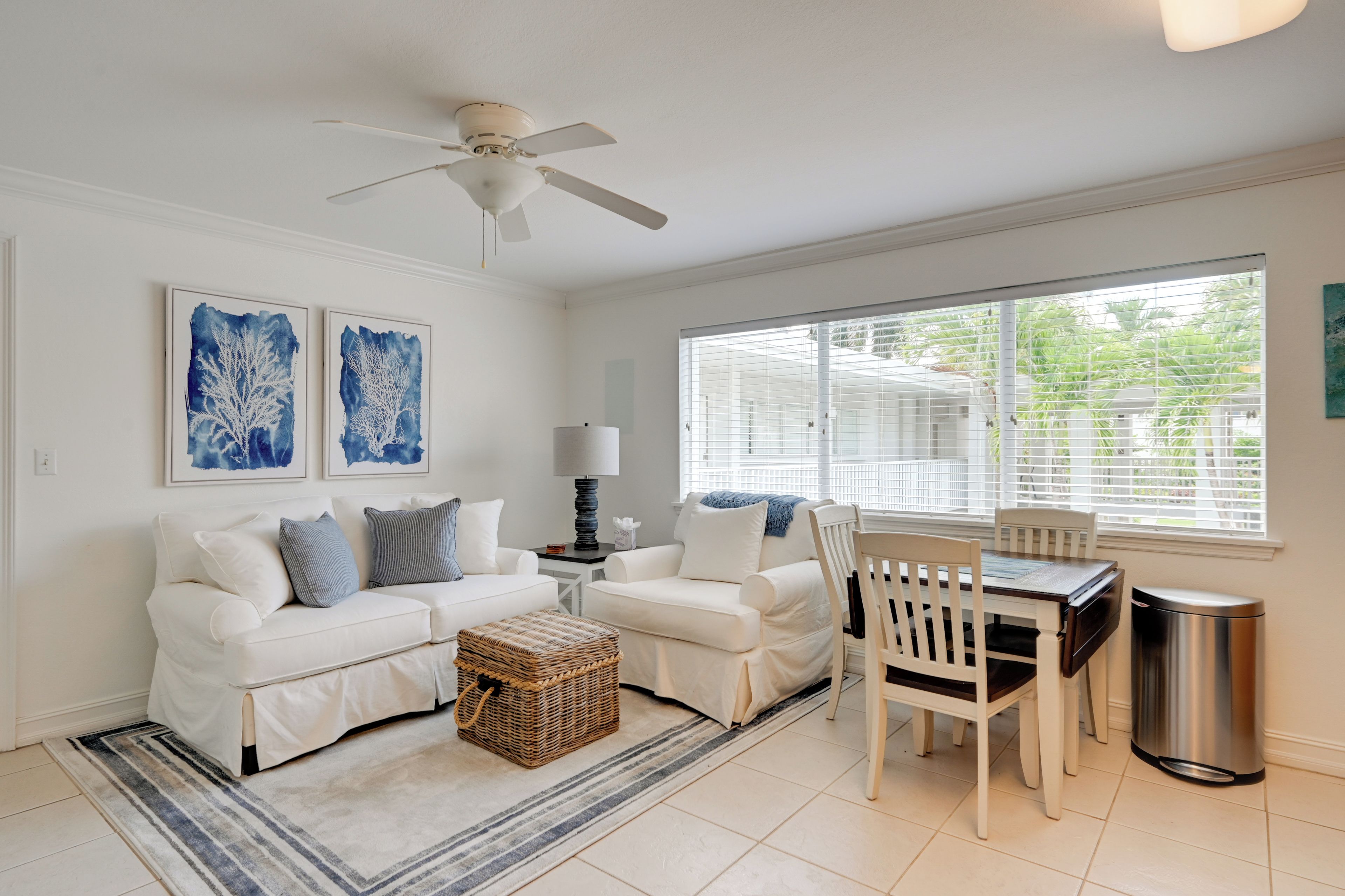 A living room with a white sofa set, a dining table with chairs, a ceiling fan, and artwork on the walls.