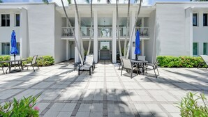 Un hotel con zona de estar al aire libre cubierta, palmeras y un edificio de dos pisos.
