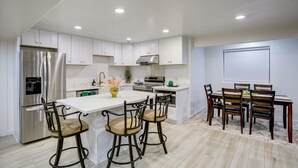 A modern kitchen with white cabinets, a stainless steel refrigerator, a dining area with a table and chairs, and a window with blinds.