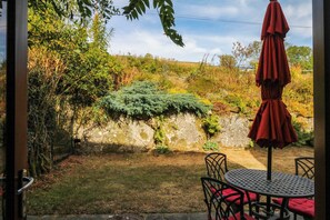 A patio with a table and chairs, an umbrella, and a view of a garden with a stone wall. - Dashel Cottage at Dockwell Farm (Newton Abbot)