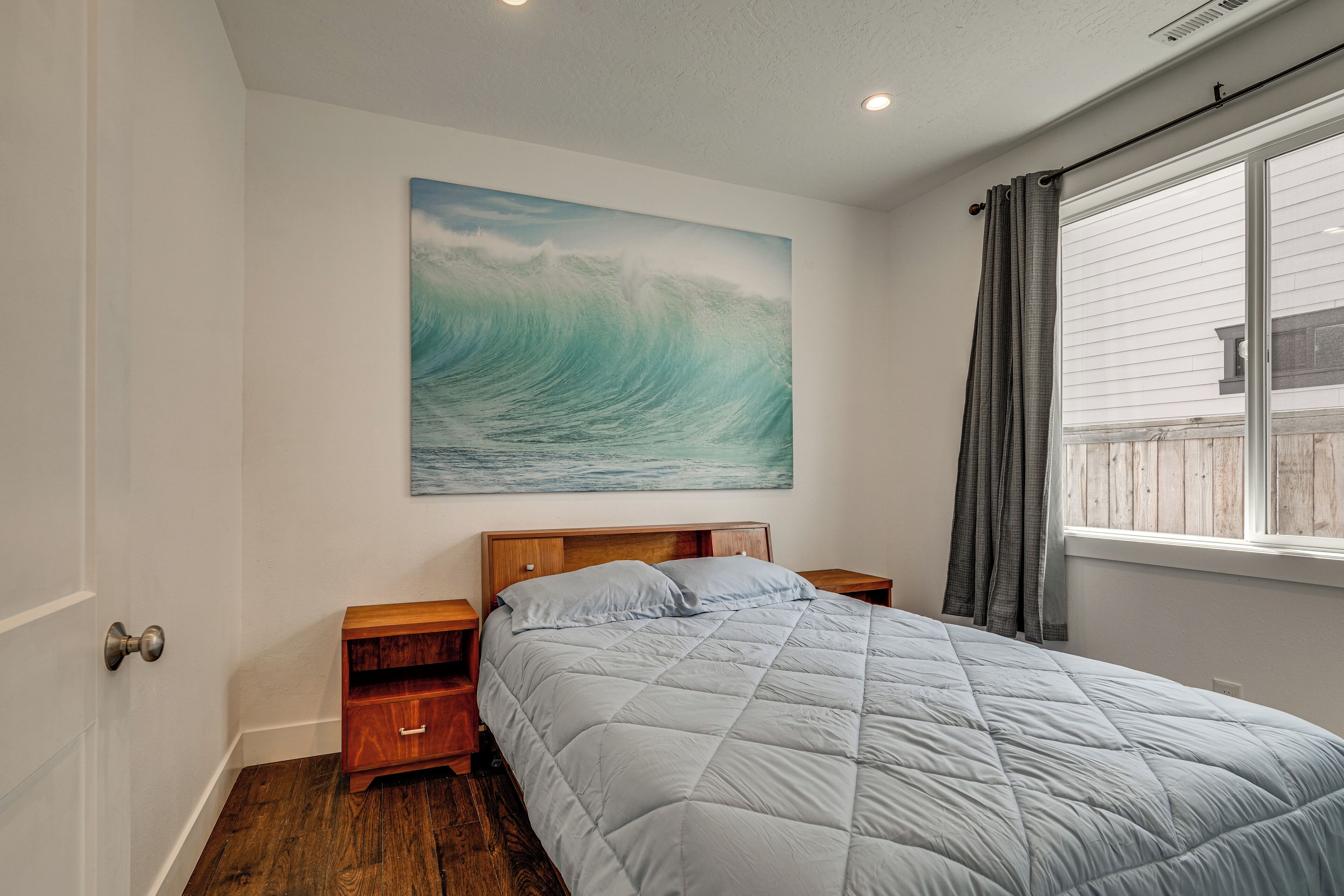 A bedroom with a large bed, wooden nightstands, a wall-mounted painting of a wave, and a window with curtains.