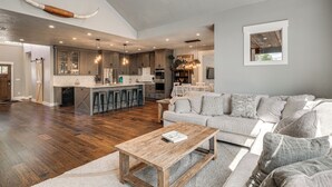 A spacious living area with a wooden coffee table, a grey sofa, and a kitchen with wooden cabinets and a large island. - Walk to Autzen Stadium: Eugene Getaway w/ Yard (Eugene)