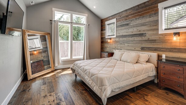 A bedroom with a large bed, wooden paneling on the walls, a window with curtains, and a nightstand with a lamp. - Walk to Autzen Stadium: Eugene Getaway w/ Yard (Eugene)