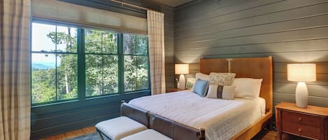 A bedroom with a large bed, two bedside tables, a bench, and a view of greenery through the window.