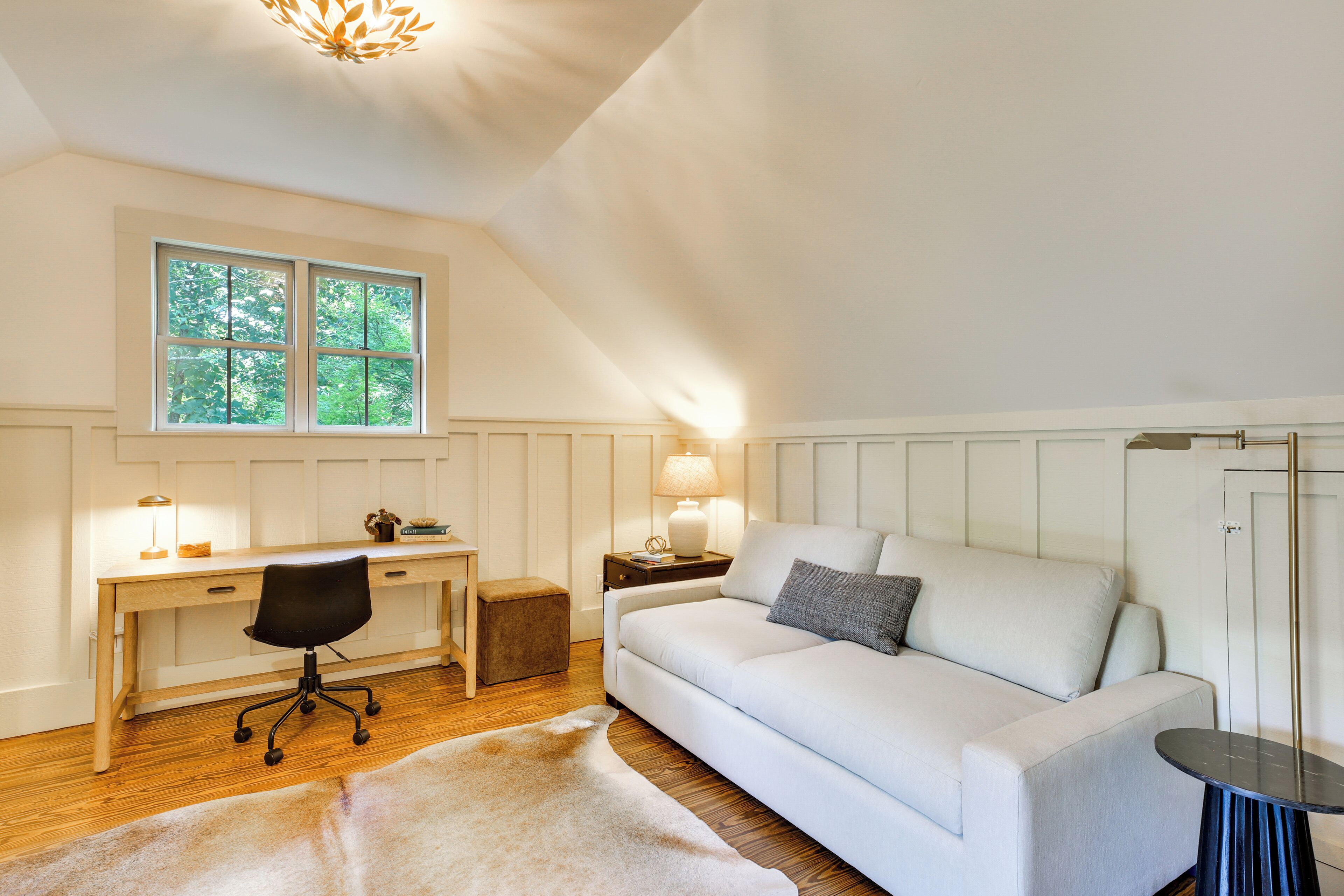 A room with a desk, chair, sofa, and a window with a view of trees.