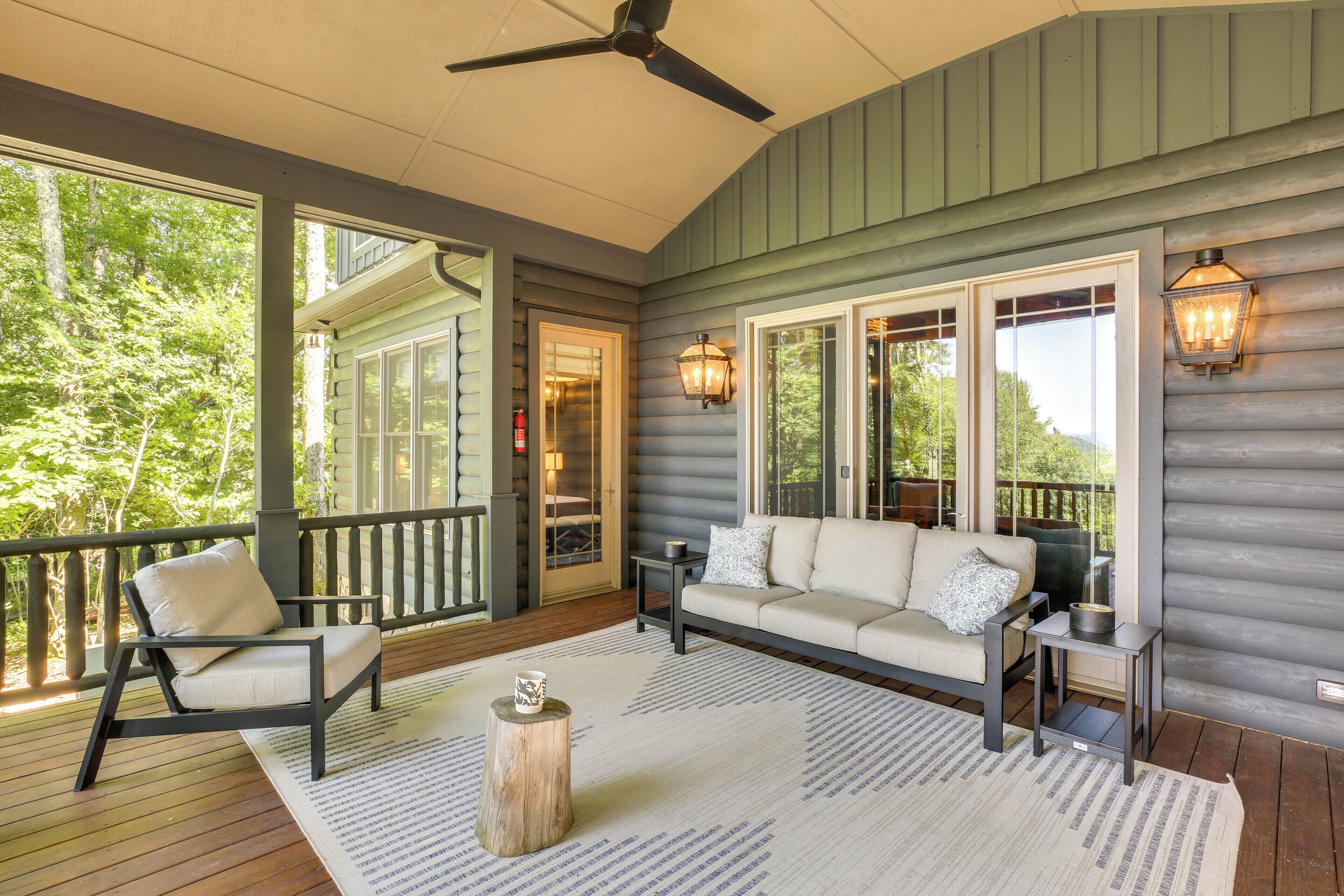 A covered porch with a sofa, chair, and table, featuring large windows and a ceiling fan.