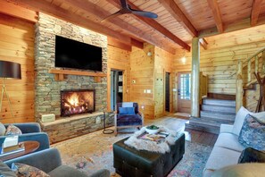 A log cabin interior with a stone fireplace, a flat-screen TV mounted above it, and a staircase leading to an upper level. - Views, Hot Tub & Fire Pit! Luxe Blue Ridge Retreat (Blowing Rock)