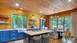 A wooden cabin with a large kitchen featuring blue cabinets, a central island with bar stools, and a dining area with a table and chairs. - Views, Hot Tub & Fire Pit! Luxe Blue Ridge Retreat (Blowing Rock)