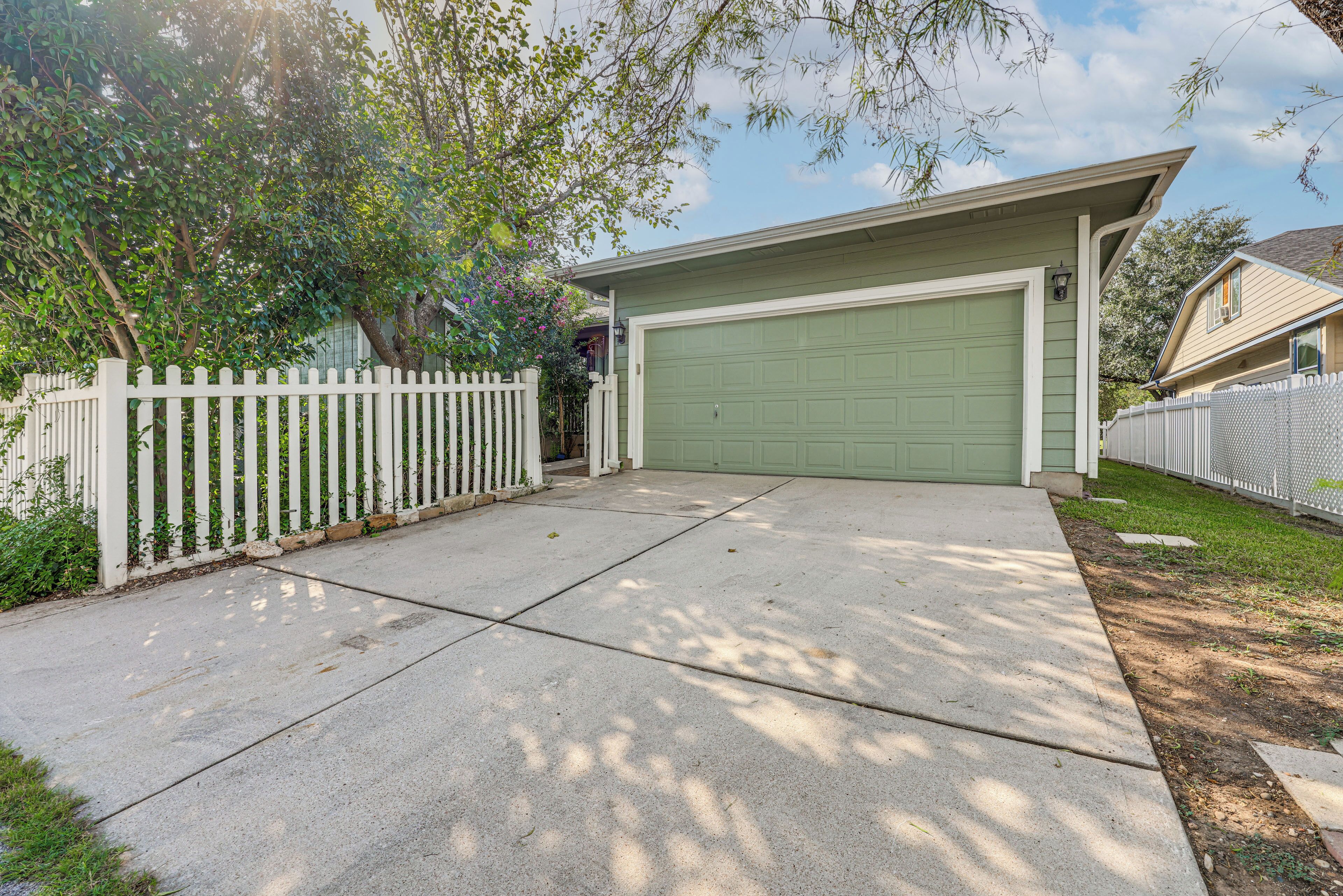 A residential garage with a green door, a white picket fence, and a tree in the background.
