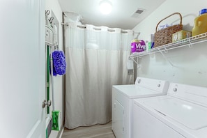 A laundry room with a washing machine, a drying rack, a shelf with cleaning supplies, and a shower curtain.