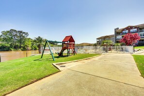 A playground with a swing set and a red playhouse, a concrete pathway, and a building with a white fence in the background. - Lake Martin Condo Getaway With Pool! (Alexander City)
