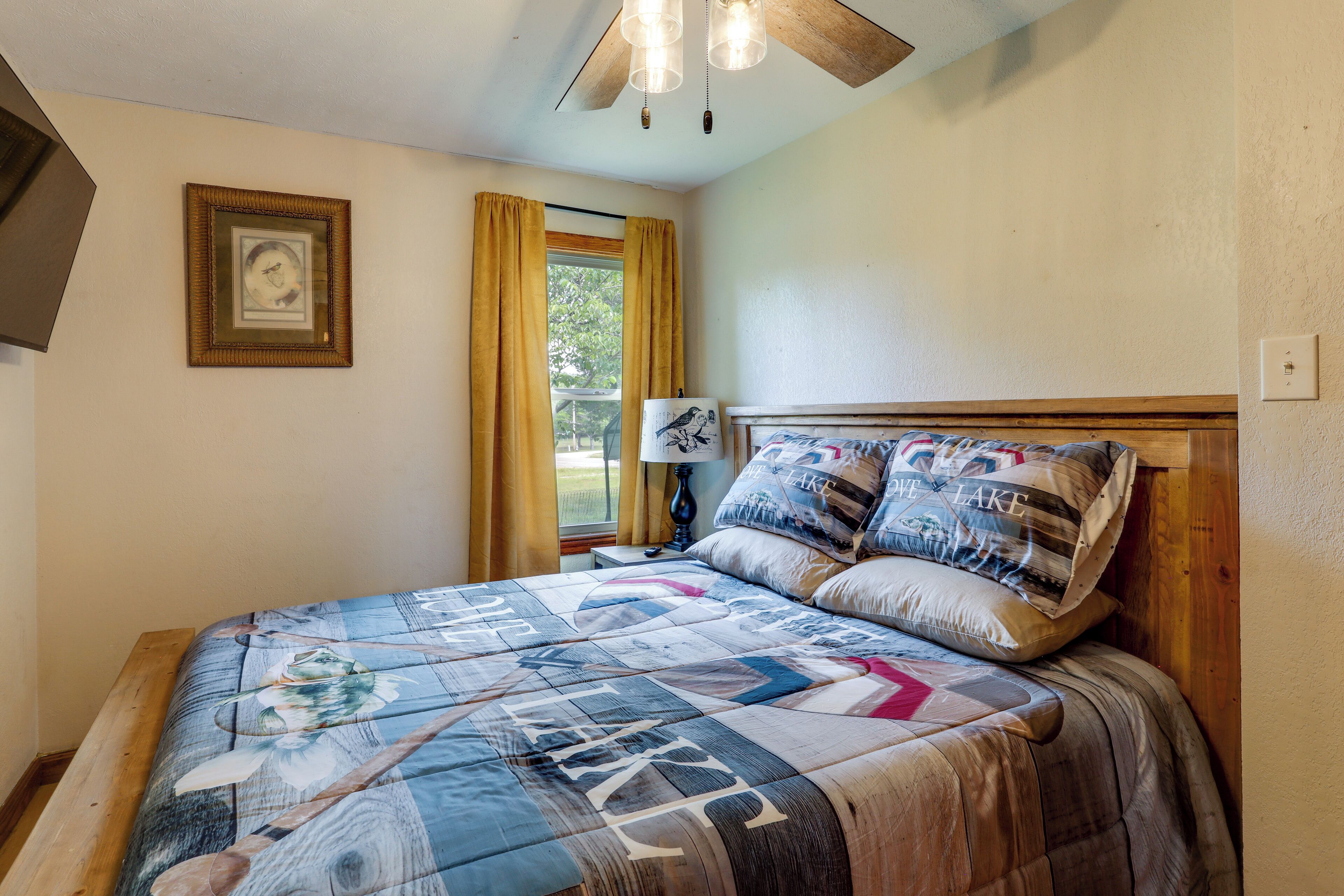 Quarto com cama, mesa de cabeceira, luminária, quadro na parede e janela com cortinas.