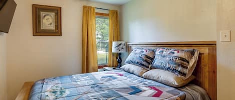 Quarto com cama, mesa de cabeceira, luminária, quadro na parede e janela com cortinas.