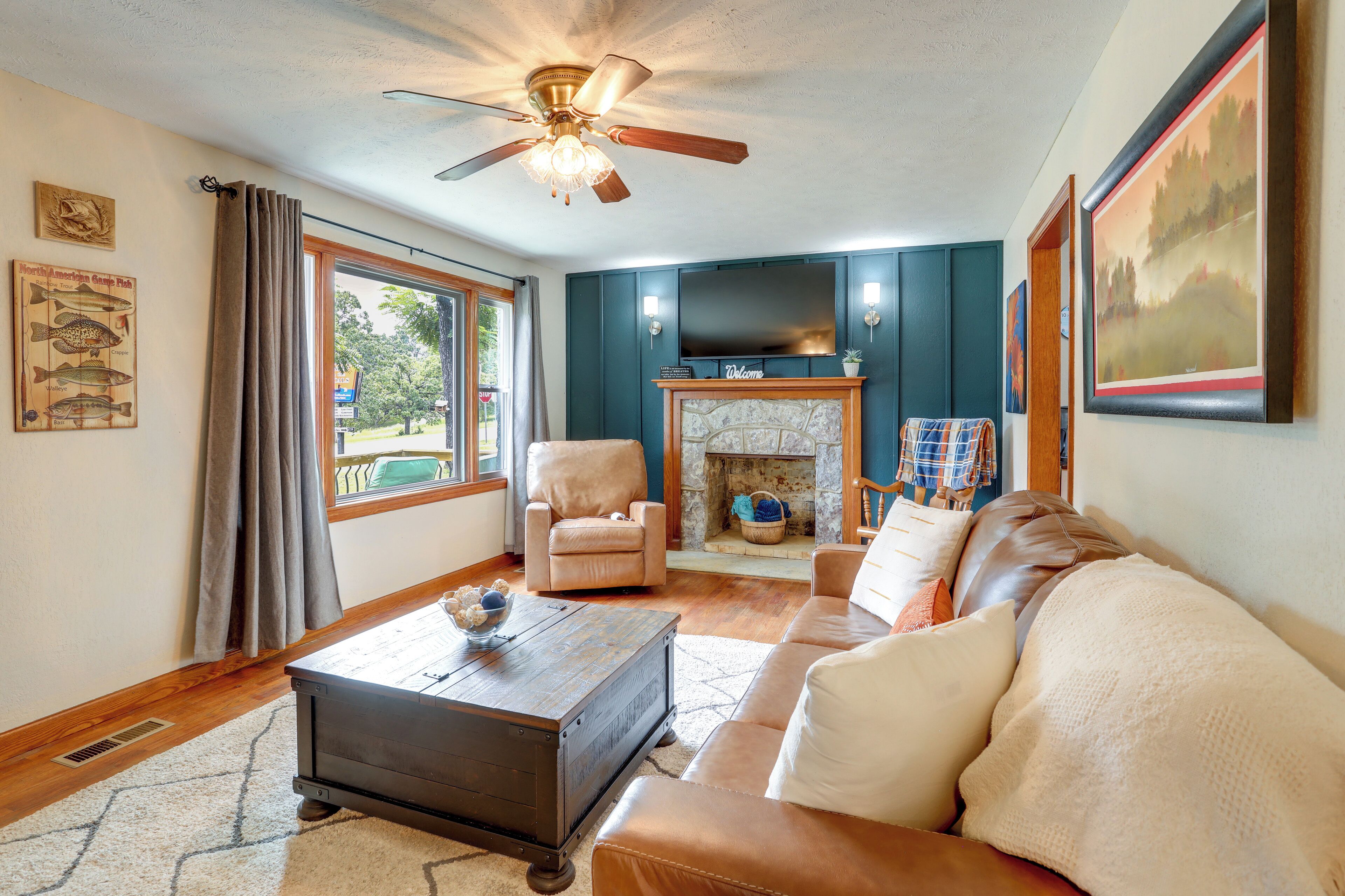 A living room with a fireplace, a television, a ceiling fan, and a large window.