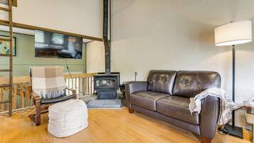 A cozy living room with a sofa, a wooden chair, a small ottoman, a fireplace, and a television mounted on the wall.