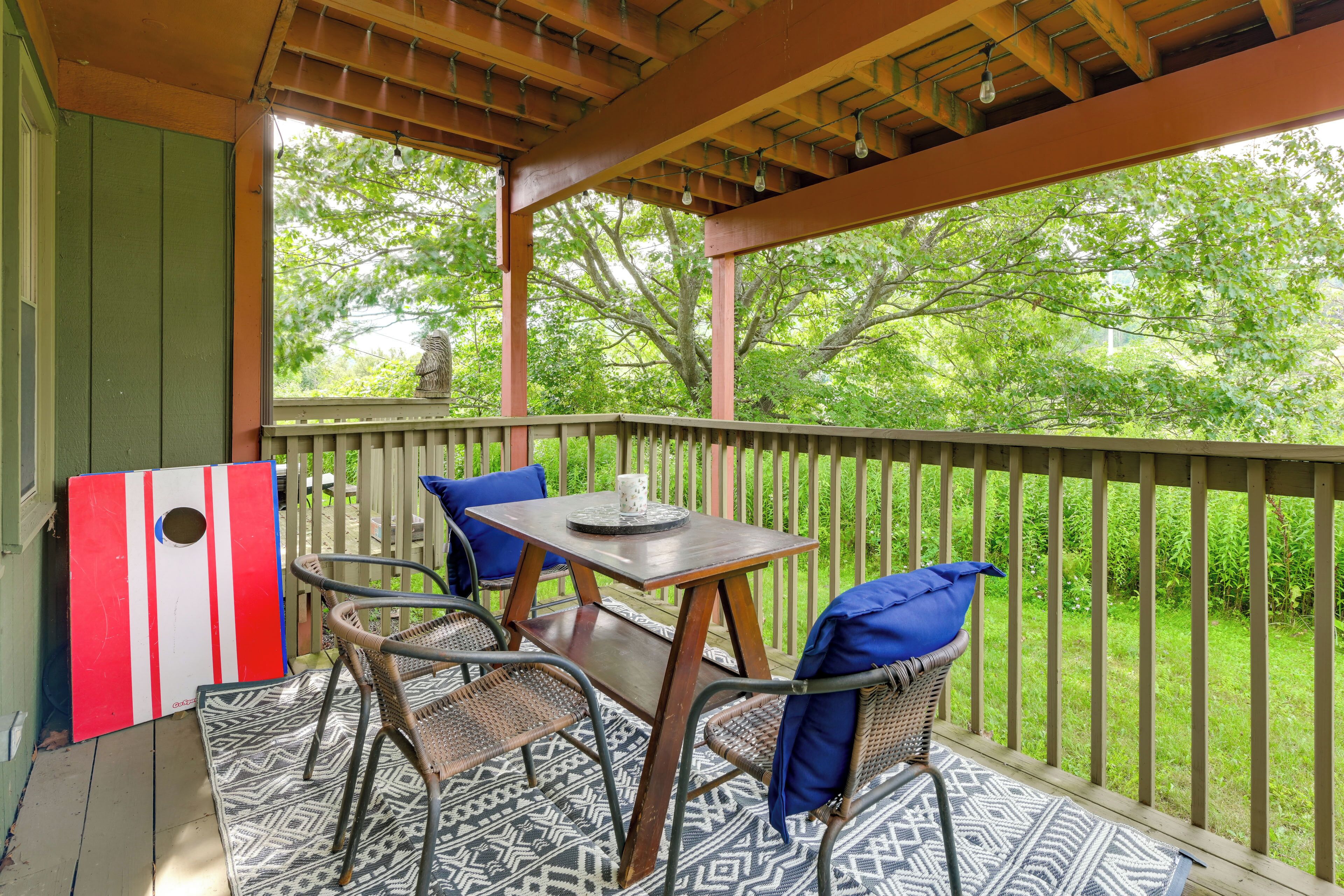 A covered outdoor seating area with a table and chairs, a decorative wall piece, and a view of greenery.