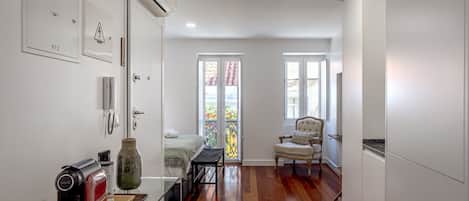A compact room with a bed, a chair, a small table with a kettle, and a window with a view of a balcony.