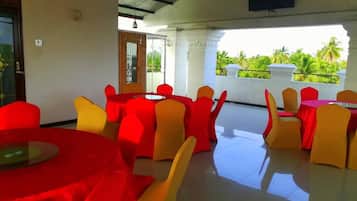 A restaurant with red and yellow chairs around tables, a flat-screen TV mounted on the wall, and a view of greenery outside.