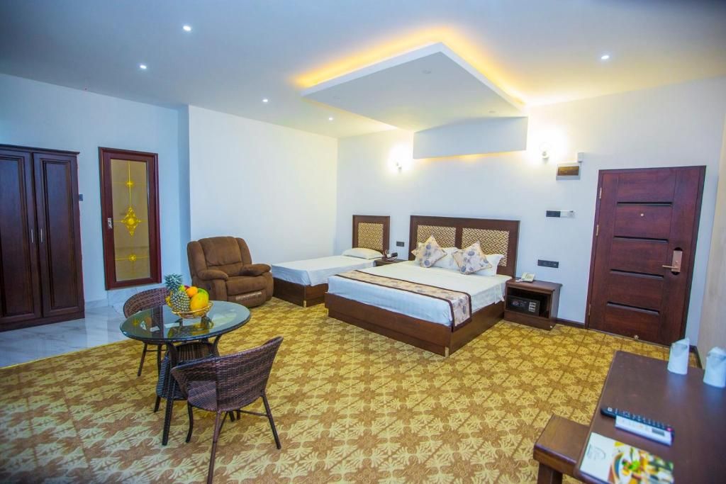 A hotel room with a bed, a desk, a chair, a small table with a fruit bowl, and a patterned carpet.