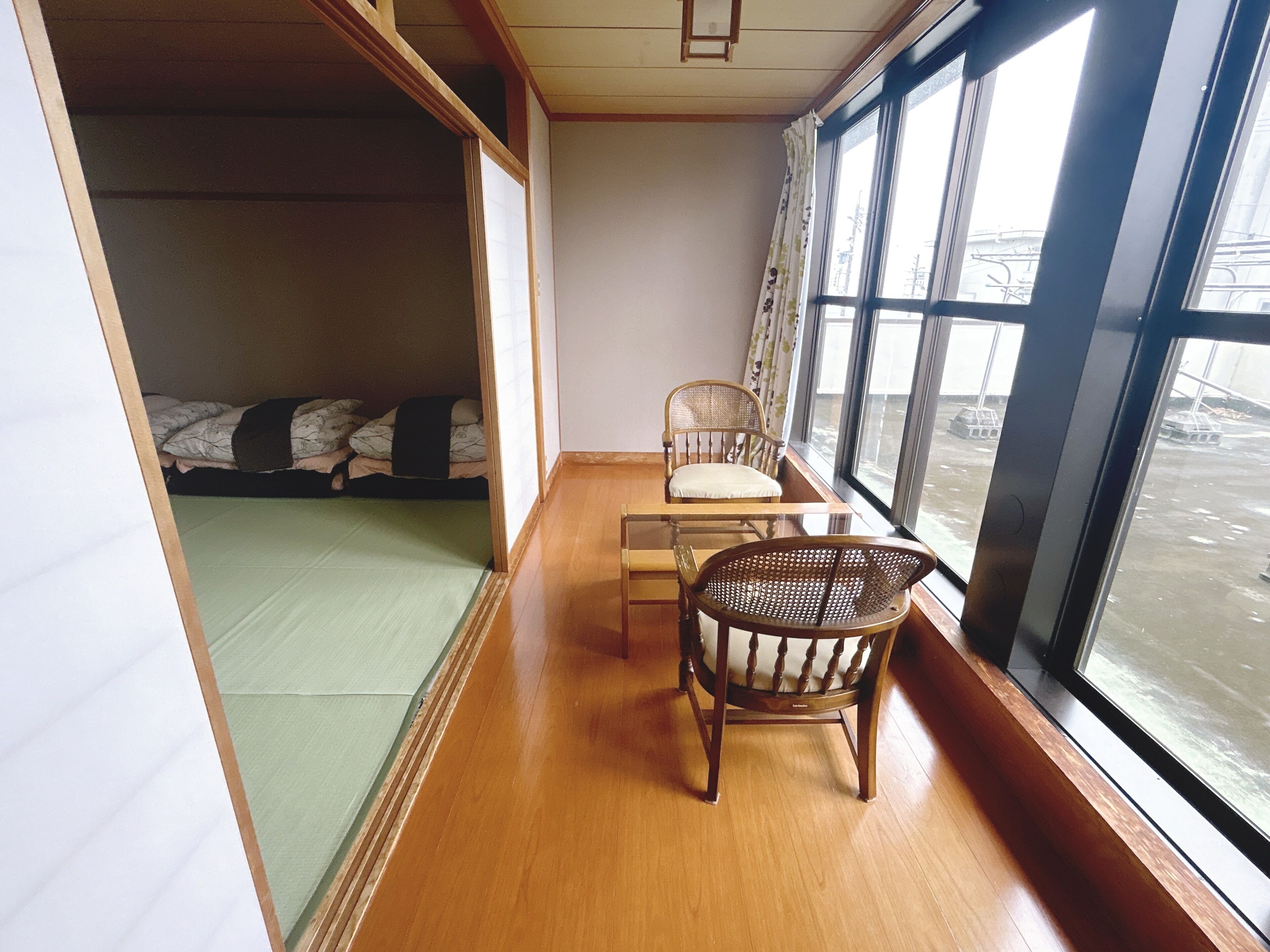 Habitación con piso de madera, una mesa con sillas, una cama con colchoneta verde y ventanas amplias con vistas al exterior.