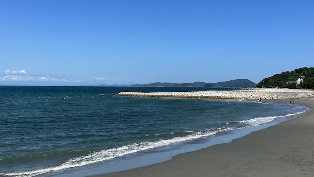 A beach with a stone breakwater, clear skies, and distant mountains.