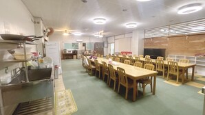 A dining area with multiple tables and chairs, a kitchen area with appliances, and a ceiling with recessed lighting.