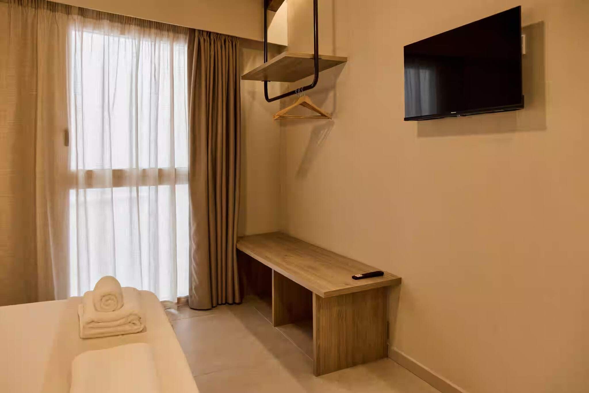 Habitación de hotel con cama, mesita de noche, control remoto, televisor montado en la pared y ventana con cortinas.