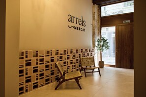 Un área de recepción con un cartel en la pared que dice 'arrels MERCAT', dos sillas de mimbre, una planta en maceta y una puerta de madera con un letrero encima.