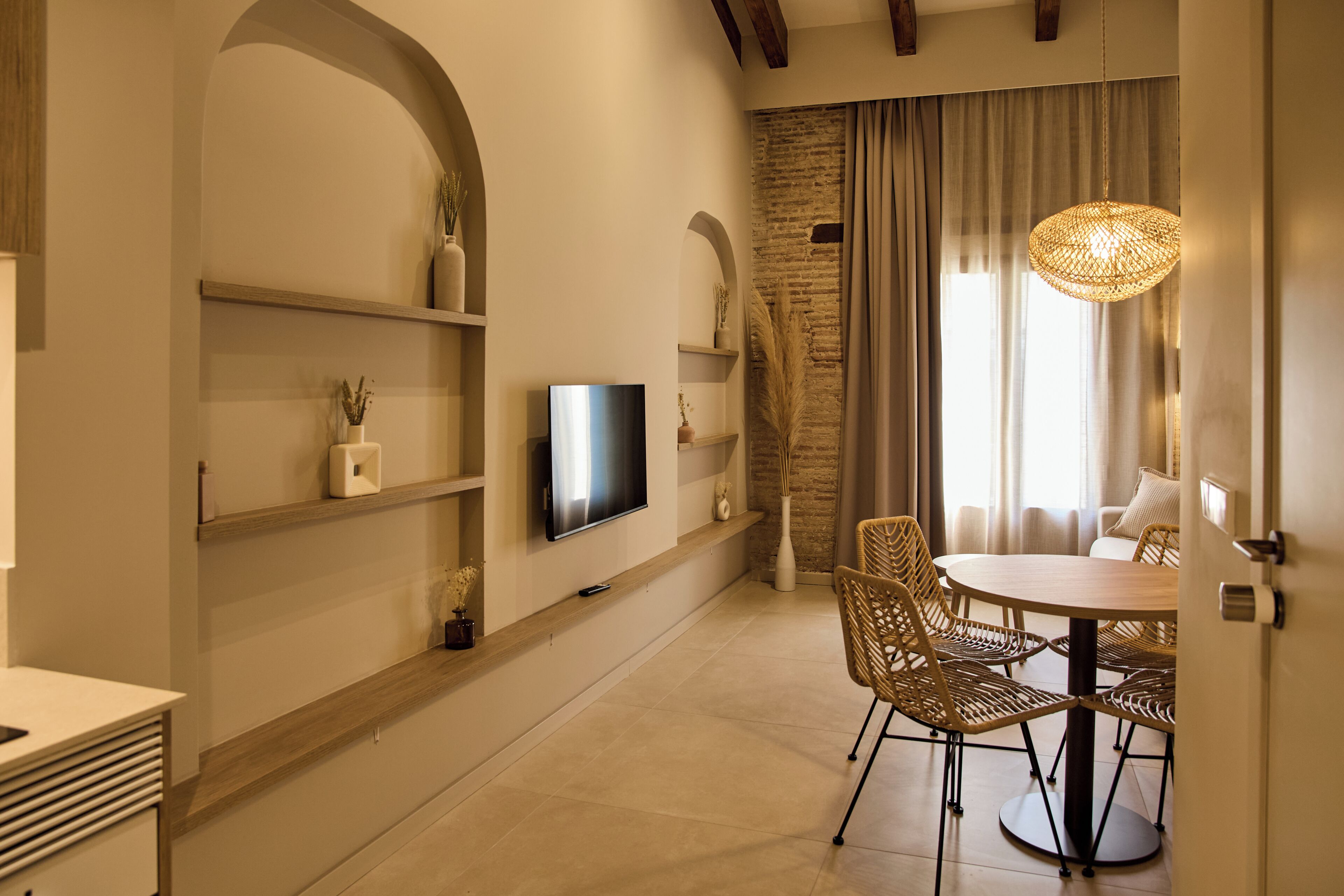 Un interior moderno con zona de comedor, televisor de pantalla plana montado en la pared y un ventanal con cortinas.