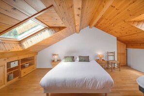 Quarto com teto inclinado, uma cama, duas mesas de cabeceira com abajures, uma cadeira e nichos embutidos.