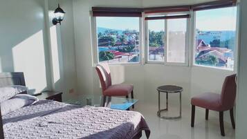 A bedroom with a bed, two chairs, a small table, and a view of a residential area through large windows.