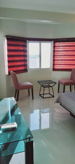 Una habitación con cama, dos sillas, una mesa de vidrio y una ventana con persianas rojas.