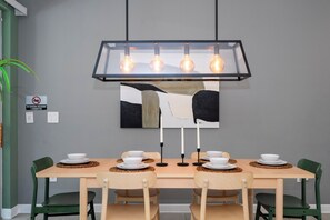 Een eetruimte met een rechthoekige tafel, zes stoelen, een moderne hanglamp en een aan de muur bevestigd schilderij.