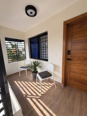 Ein Zimmer mit Holztür, zwei weißen Stühlen, einer Topfpflanze und einem Fenster mit Metallgitter.