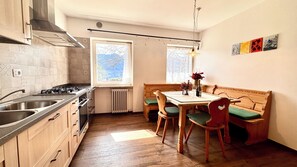A kitchen with a stove, sink, and wooden cabinets. There is a dining table with chairs and a bench. - Villa ai Noci (Carano)