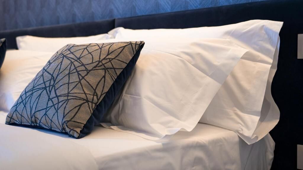 A bed with white linens and a patterned pillow.