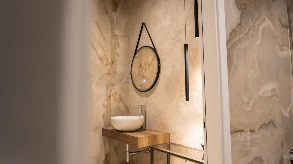 A bathroom with a marble wall, a wooden vanity, a round mirror, and a white sink.
