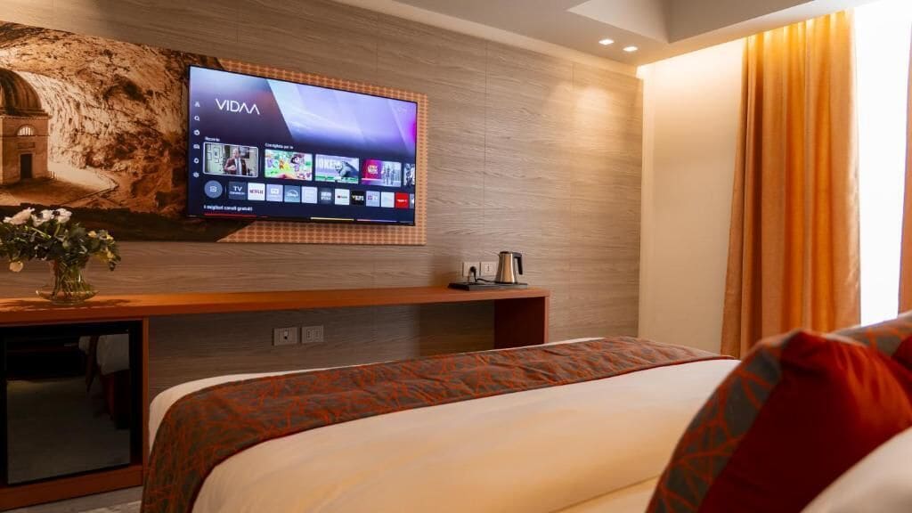 Ein Hotelzimmer mit einem großen Flachbildfernseher an der Wand, einem Bett mit gemusterten Betttextilien, einem kleinen Tisch mit einem Wasserkocher und einer dekorativen Vase mit Blumen.