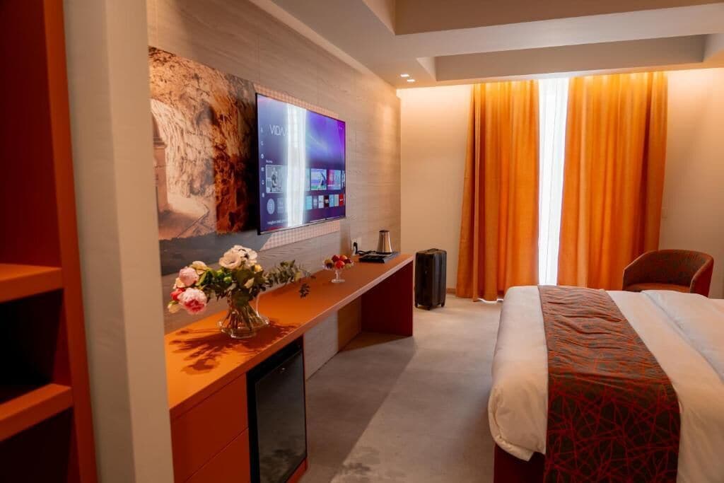 A hotel room with a bed, a desk with a flat-screen TV, a vase of flowers, and a window with curtains.