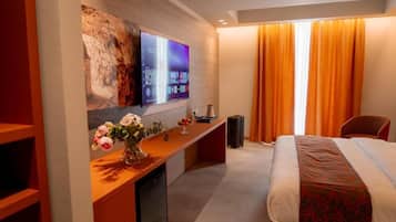 A hotel room with a bed, a desk with a flat-screen TV, a vase of flowers, and a window with curtains.