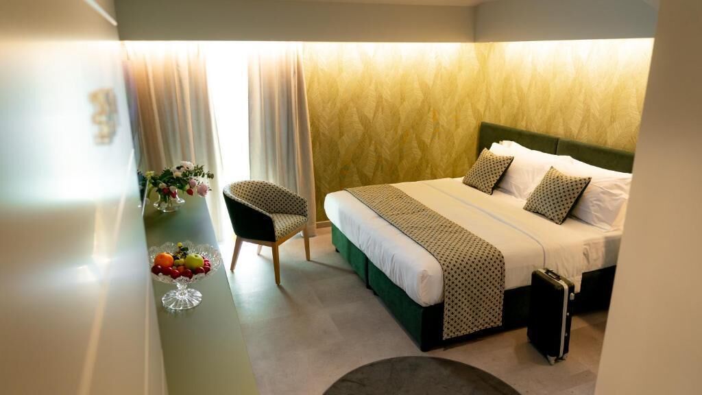 A hotel room with a bed, a chair, a small table with fruit, and a suitcase.