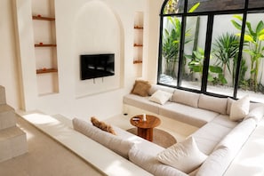 A modern living room with a sectional sofa, a flat-screen TV mounted on the wall, and a large window with plants. - 2BR Private Pool Villa Walk to Pererenan Beach and Cafes (Mengwi)