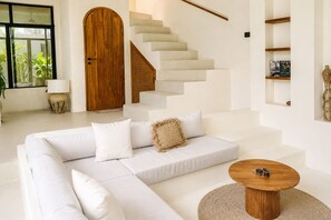 A modern living room with a white sofa, a round wooden coffee table, and a staircase with a wooden door. - 2BR Private Pool Villa Walk to Pererenan Beach and Cafes (Mengwi)