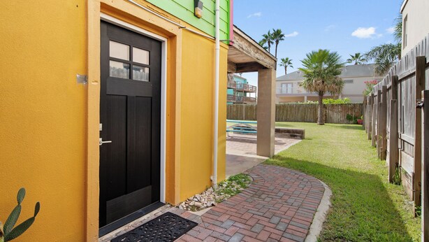 A yellow building with a black door, a brick pathway, and a small grassy area with a wooden fence. - Spacious Beach House w/Fenced Yard, Firepit, Patio & Walk to Beach! (South Padre Island)