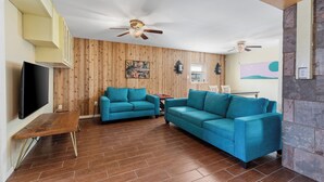 A living room with two teal sofas, a wooden floor, a ceiling fan, and a TV on the left wall. - Spacious Beach House w/Fenced Yard, Firepit, Patio & Walk to Beach! (South Padre Island)